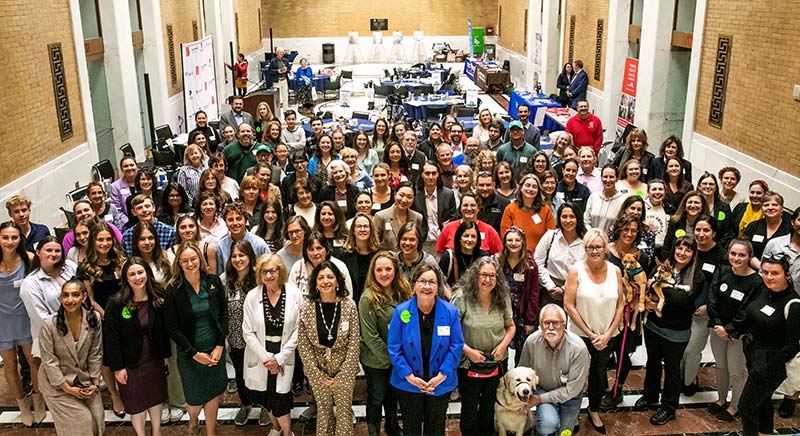A large, diverse group of people, including a person with a dog, smiling in a spacious hall with tables.