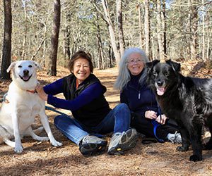 Two smiling people sit on a forest path with two dogs, one black and one white. The scene is cheerful, with sunlight filtering through trees.