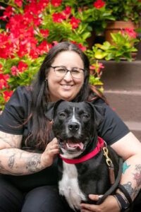 A woman with long dark hair and glasses sits on outdoor steps, smiling beside a black dog with a white chest and a red collar. Red flowers and green plants are in the background.