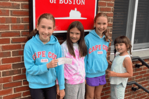 Four young girls smile in front of a brick wall. Two wearing blue Cape Cod sweatshirts, one holds a check, with a sign reading 