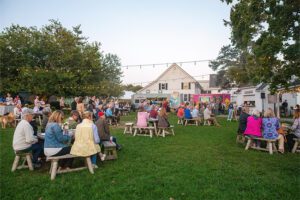 People gather at picnic tables on a grassy lawn under string lights, enjoying a lively outdoor event near charming rustic buildings at dusk.