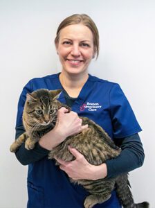 A veterinary professional in a blue uniform gently holds a tabby cat, smiling warmly against a plain background, conveying care and compassion.