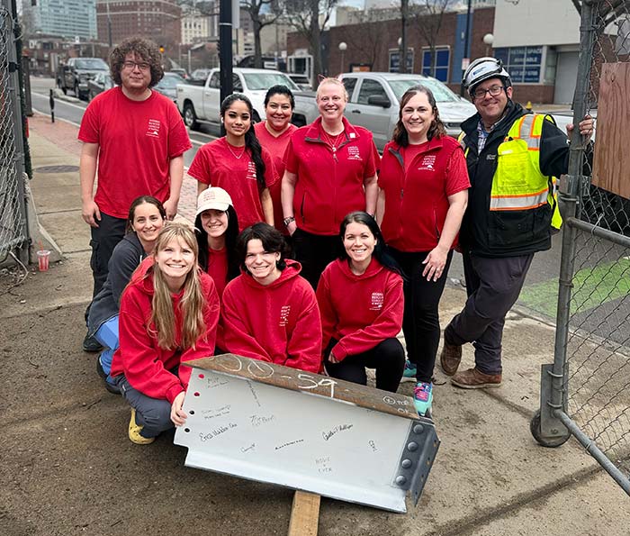 A group of eleven people, mostly in red shirts, pose cheerfully on a construction site. They gather around a signed metal beam.