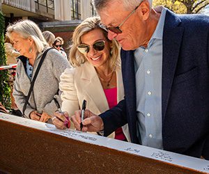A couple in sunglasses is cheerfully signing a large beam outdoors. They are surrounded by people, with a sunny and festive atmosphere.