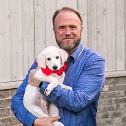 A bearded man in a blue shirt holds a white puppy with a red bandana, against a rustic wooden background.