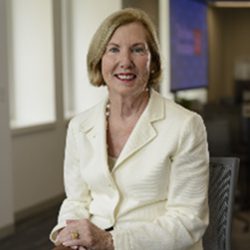 A woman in a white blazer sits smiling in an office. The room is softly lit, creating a warm and professional atmosphere.