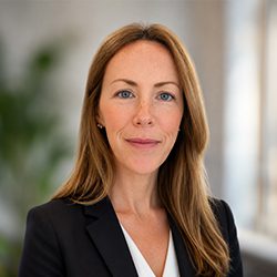 A woman with long brown hair, wearing a black blazer and white blouse, poses confidently. The background is softly blurred with a hint of greenery.