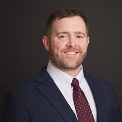 A man in a blue suit and maroon tie smiles warmly against a dark background. He has short hair and a neatly trimmed beard.